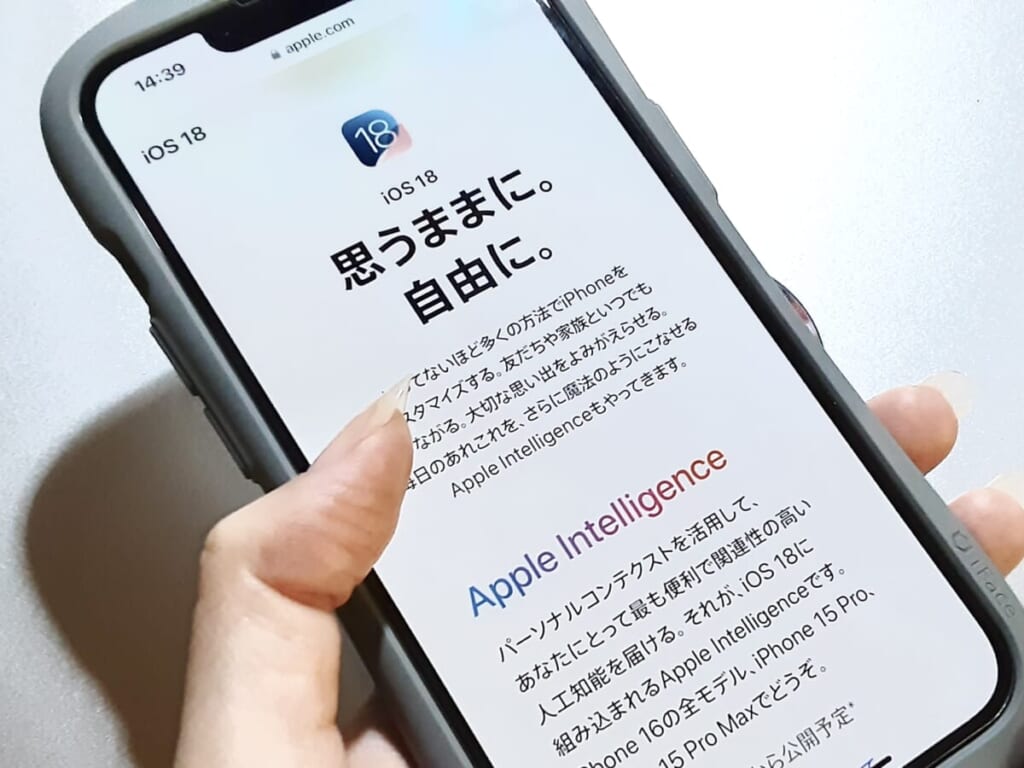 iPhoneを手に持ち、Apple公式サイト「iOS 18正式リリースページ」を紹介する画像