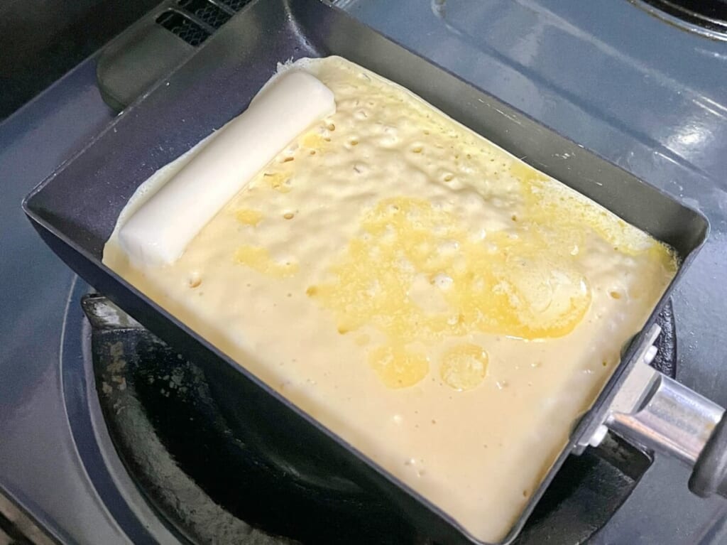 卵焼き器にチーズドッグの生地とマーガリンを入れて焼き、端にさけるチーズを置いた様子