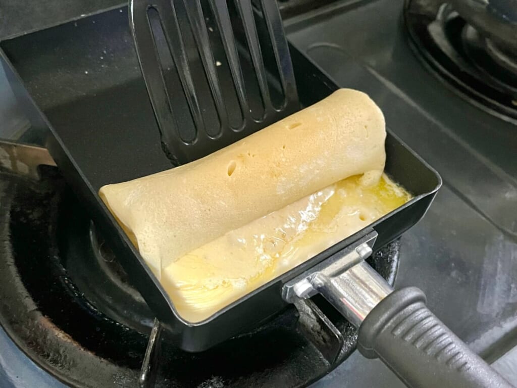 卵焼き器にチーズドッグの生地とマーガリンを入れて焼き、端にさけるチーズを置いて内側に向かって巻いている様子